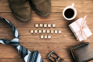 Flat lay of Father's Day gift items: shoes, coffee, gift box, striped tie, wallet, belt on wood.