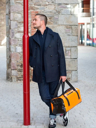 Man in navy pea coat, dark jeans, and sneakers holding a bright orange duffel bag outdoors.