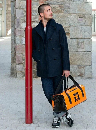 Man in navy pea coat and jeans holding orange duffel bag, city street background