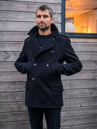 Man wearing a navy wool pea coat with double-breasted buttons standing outside by a wooden wall