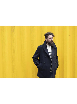 Man in navy wool pea coat and striped shirt standing against yellow wall