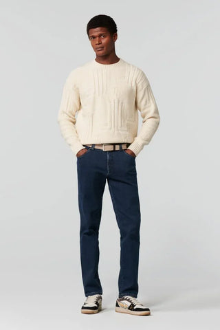 Man wearing dark blue Meyer jeans, cream sweater, and sneakers, standing against plain background