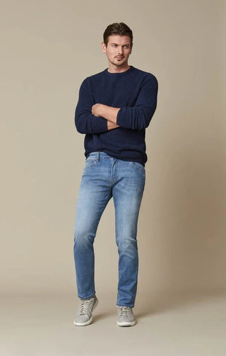 Man wearing light blue slim-fit jeans, navy sweater, and grey sneakers on beige background