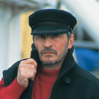 Man wearing black wool captain cap with braid detailing, red sweater, zip-up vest outdoors