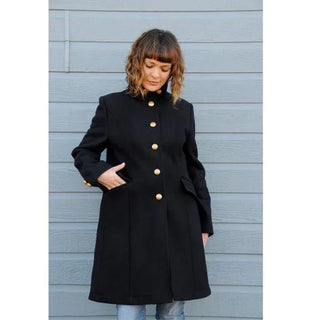 Woman in long black wool coat with gold buttons standing against gray wall