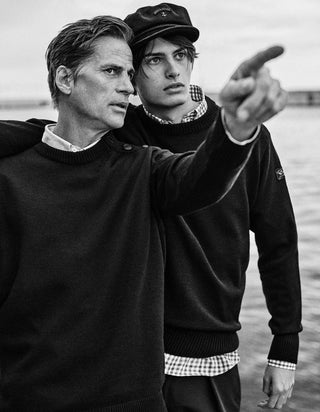 Two men in dark sweaters and a wool captain's cap by the water, one pointing forward.