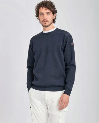 Man wearing navy crewneck sweater with shoulder patches and white pants, standing indoors
