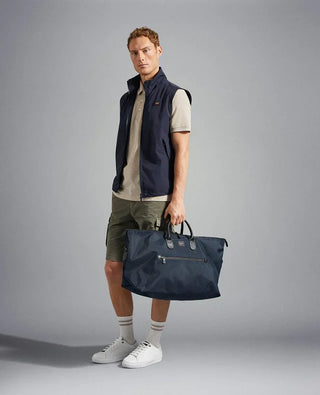 Man wearing a navy nylon sleeveless vest, beige polo shirt, green shorts, holding a large navy duffel bag