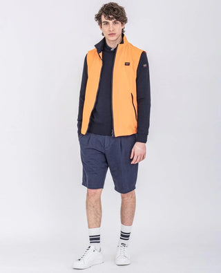 Man wearing orange sleeveless nylon vest, navy sweatshirt, navy shorts, white sneakers